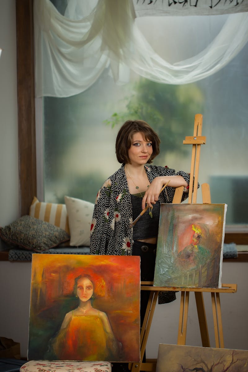 A woman artist stands by her easel displaying two vibrant portrait paintings.